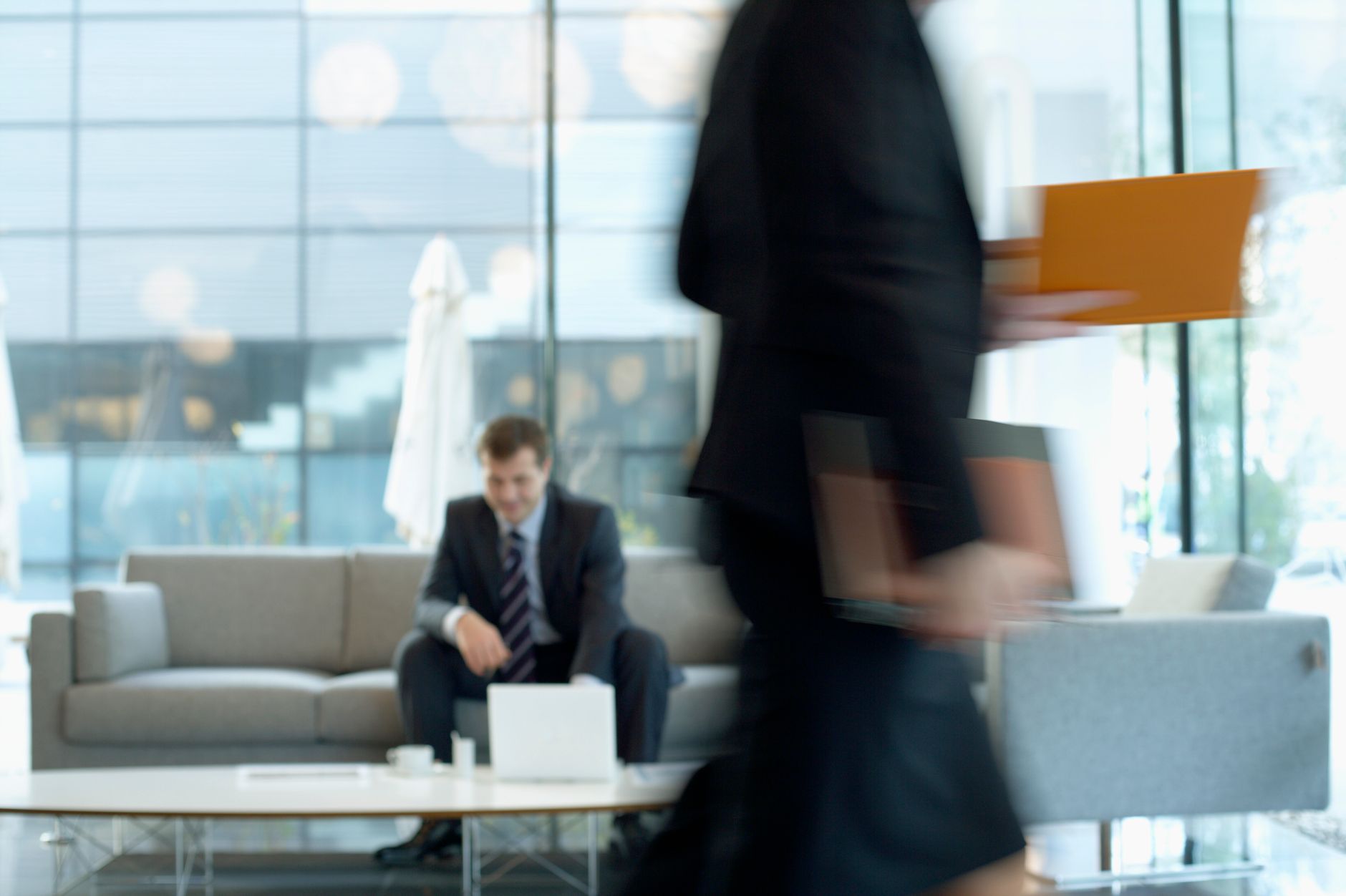 Businessman sitting in modern office lounge using a laptop while colleague walks past, creating dynamic blur. — A Touch of Th