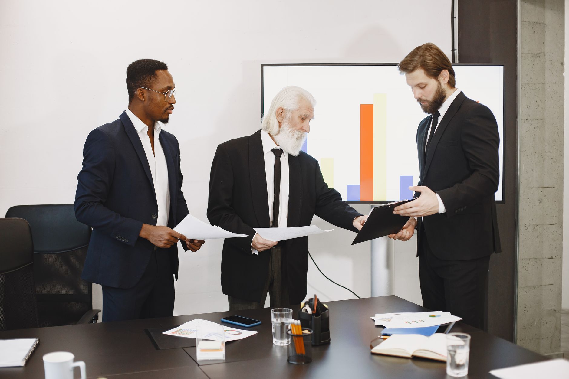 Three businessmen discussing documents in a conference setting with a presentation screen. — Droopy Eyelids: Tips For Bright