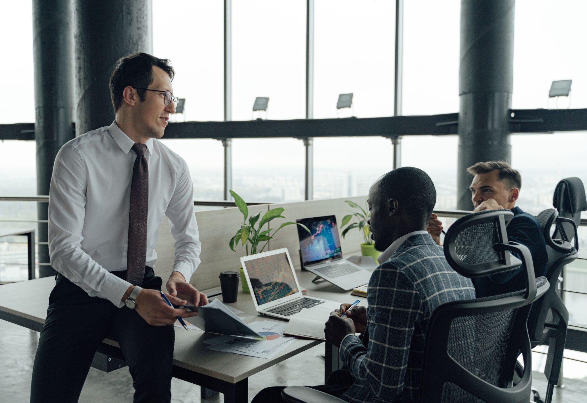 Three men engaged in a business meeting in a modern office setting with laptops and documents. — Find the Best Way to Buy Che