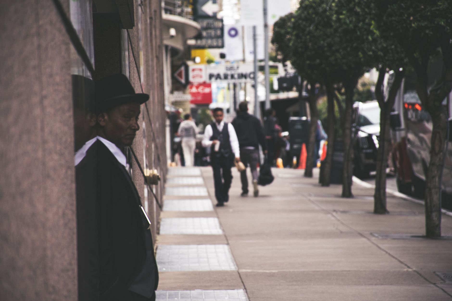 A man in a suit and hat leans against a city building, observing pedestrians. — Implementing Lasting Change: The Benefits of