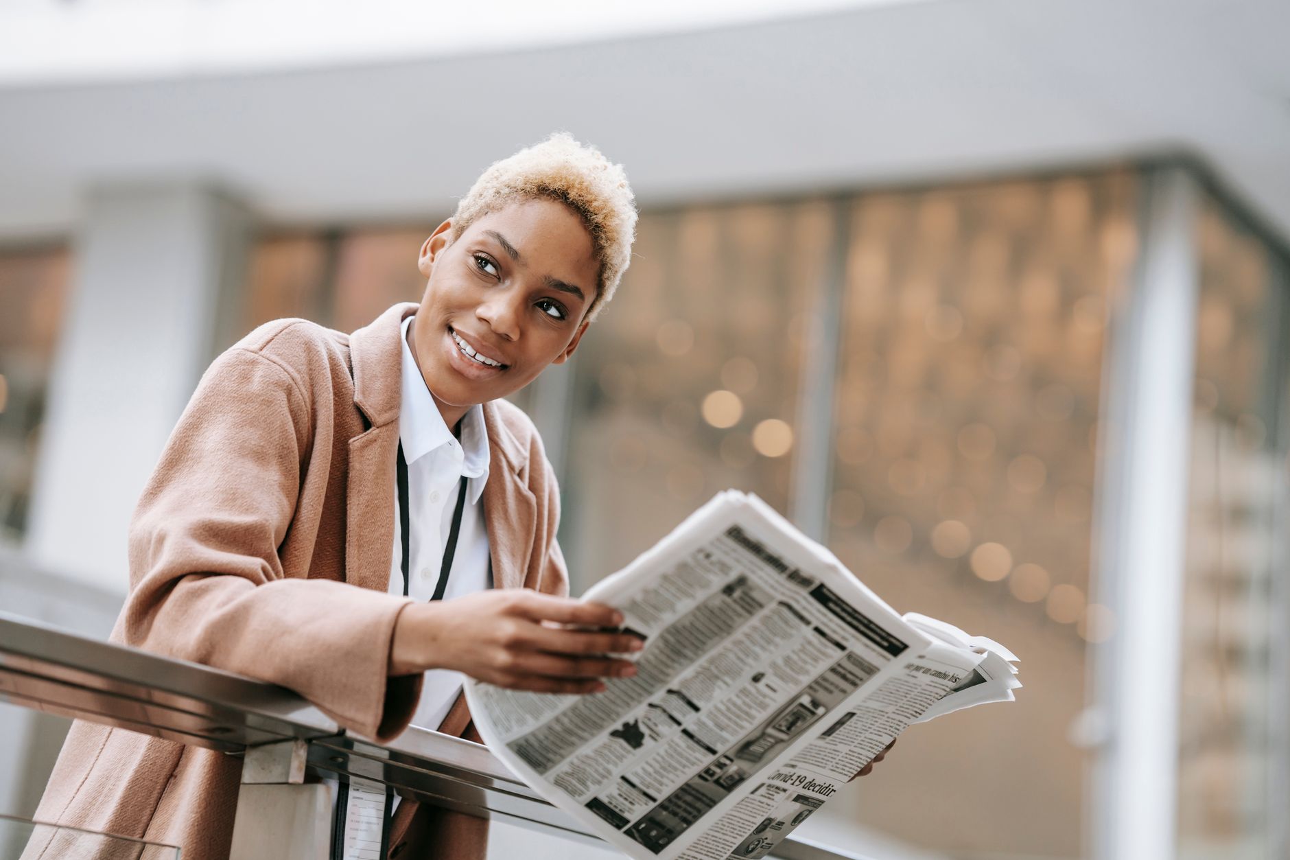 From below of positive young African American female entrepreneur in elegant brown coat reading newspaper leaning on railing