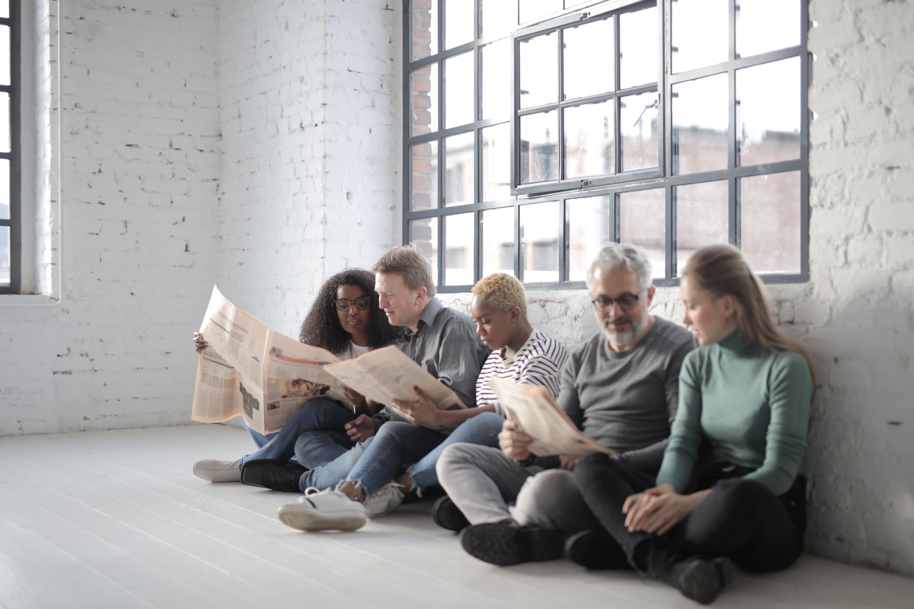 Group of diverse coworkers sitting with crossed legs on ceramic floor near fenced window in afternoon while reading newspaper