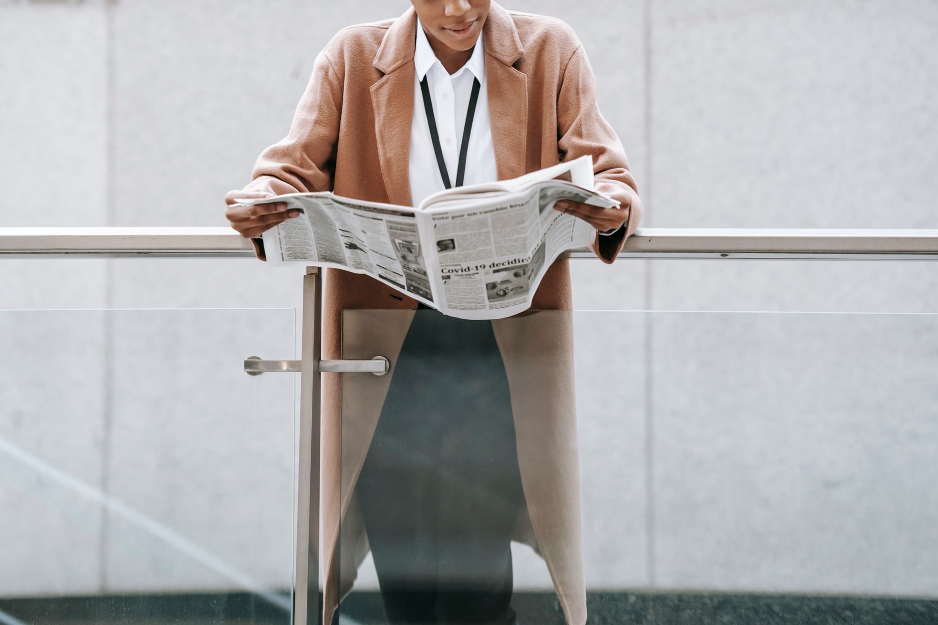 Crop content African American businesswoman in formal clothes reading fresh newspaper and leaning on glass railing of contemp