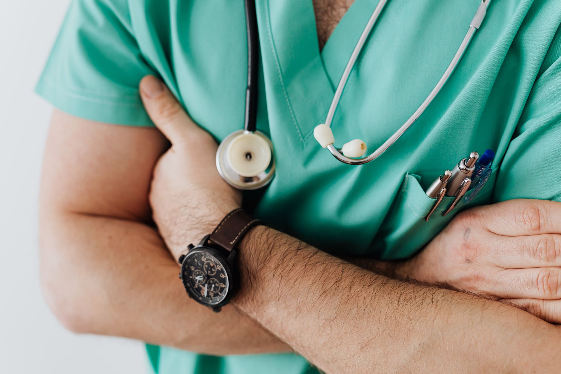 Unrecognizable man wearing medical uniform with stethoscope and pens standing in hospital with crossed arms during work in da