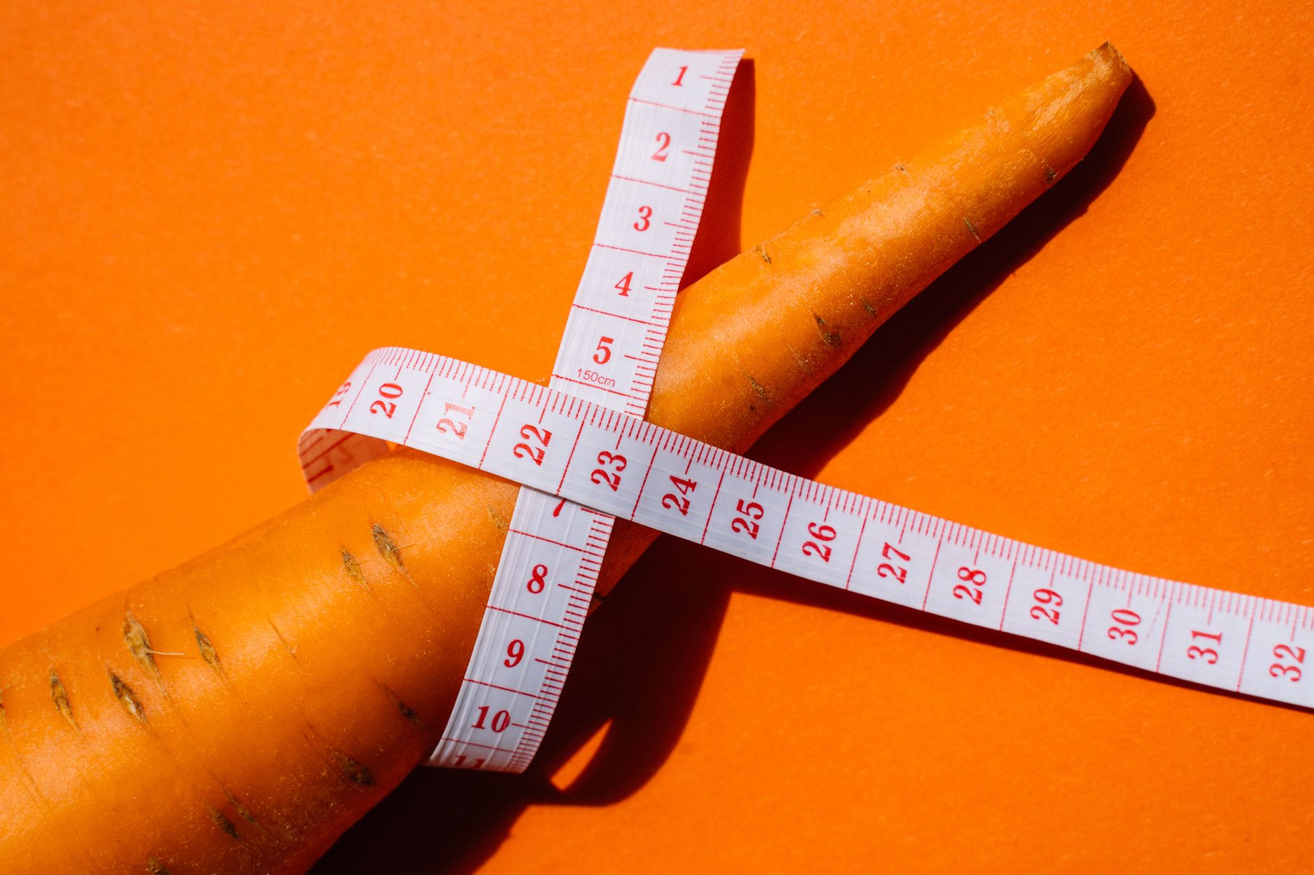 Close-up shot of a carrot wrapped with a measuring tape on a vibrant orange background, symbolizing healthy eating. — Today's