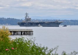 A large aircraft carrier navigating through a calm body of water with distant hills in the background.