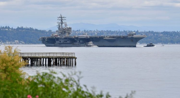 A large aircraft carrier navigating through a calm body of water with distant hills in the background.