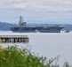 A large aircraft carrier navigating through a calm body of water with distant hills in the background.